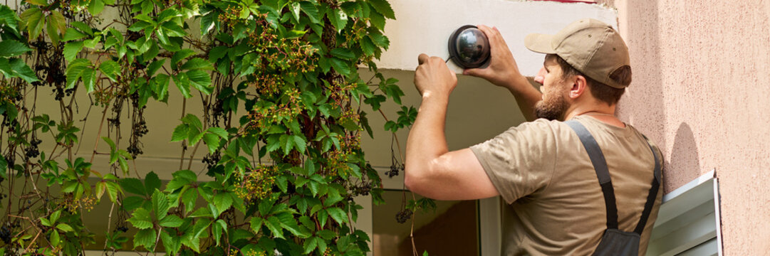Caucasian young adult male technician installing outdoor security camera on building wall. Image represents home security services and smart surveillance system installation