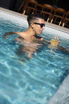 Man is standing waist-deep in clear blue pool holding cocktail with sunglasses, deck chairs behind