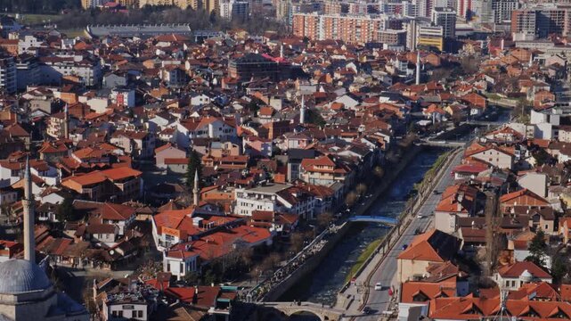 Aerial tilt up shot over Prizren city in Kosovo revealing Sinan Pasha Mosque, Lumbardhi river, traditional red roofs, and snow capped mountain peaks in the background under clear blue sky.