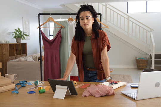 Woman leaning on wooden table, wearing olive green top and rust shirt, arranging fabric and tablet