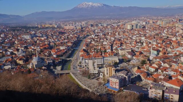 Wide panning aerial shot of Prizren city in Kosovo featuring traditional red tile roofs, Ottoman mosques along the river, and snow capped mountains in the background under a clear sky.