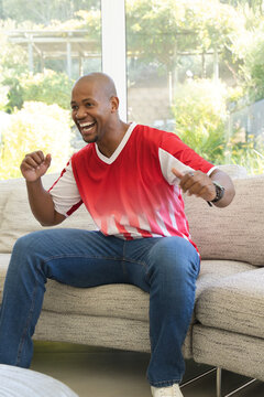 African American man sitting on sofa in living room, cheering near large window, wearing jersey