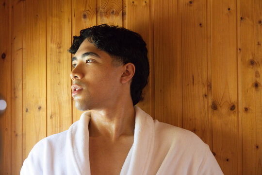 Asian man standing in wood-paneled room wearing white bathrobe, looking left with light reflection