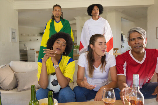 Diverse friends wearing jerseys watching soccer match at home, holding soccer ball and beer bottles