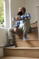 Man sitting on wooden stairs by large window at home, holding ceramic mug, wearing plaid overshirt © wavebreak3