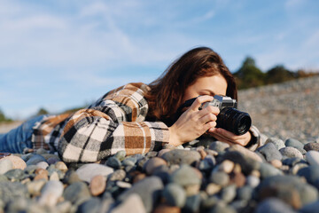 Fototapeta premium Woman photographer lying on rocky beach, aiming a camera at the horizon for landscape shots, wearing a plaid jacket, clear sky, focused expression, and a casual outdoor photography session.