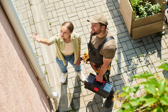 Caucasian woman pointing at gutter while talking to male contractor holding toolbox. High angle shot showing home maintenance consultation for repair service marketing