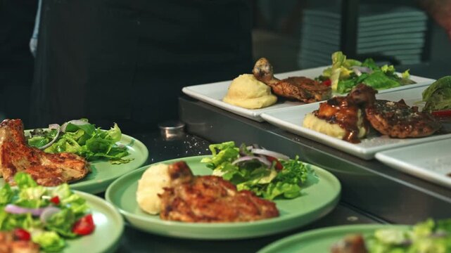 Medium tracking shot of a kitchen team garnishing, ladling sauce, and picking up finished rectangular chicken chop plates from a stainless steel pass.