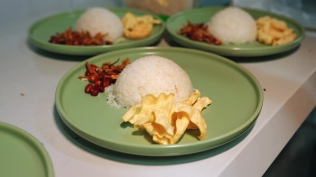 Medium tracking shot of a person wearing black gloves placing crispy crackers onto green plates lined up with coconut rice, anchovies, and peanuts on a kitchen counter.