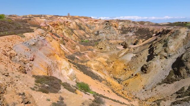 Parys Mountain Copper Mine, Amlwych, Anglesey, Wales, UK. Drone flight over cliff edge and over deep main mineralised crater towards distant derelict hilltop windmill.