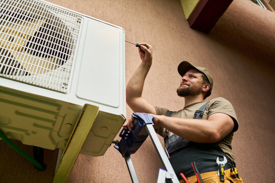 Caucasian male technician repairing outdoor air conditioning unit with screwdriver. Image suitable for HVAC maintenance services and home repair advertising, low angle view