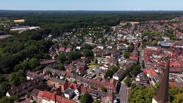 Tall church spire rises above red-roofed old town buildings in Duelmen, Germany, dense residential streets stretch toward distant wind turbines beyond forested countryside. Aerial drone approaching