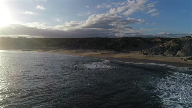 Drone pulls back and turns at Playa El Rinconcito, starting with a view down the beach toward the sun and pulling out over the ocean away from the light to reveal more of the beach and dunes to the ri