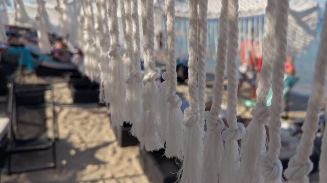 White fringe tassels sway gently in the breeze at a beach club while people relax and sunbathe near the shore 4K