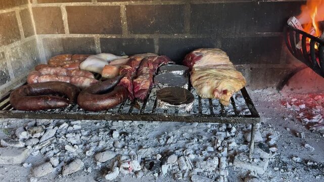 Authentic Argentine asado with beef, blood sausage, sausages, chicken and vegetables cooking over hot wood embers inside an open fireplace, panning close-up