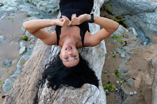 Smiling woman lying upside down on driftwood at the beach, creating a joyful and relaxed summer vibe for lifestyle and wellness concepts.