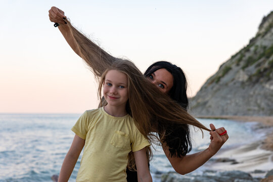 Smiling mother and daughter enjoying playful moments on a scenic beach during sunset, with mountains and calm ocean in the background.