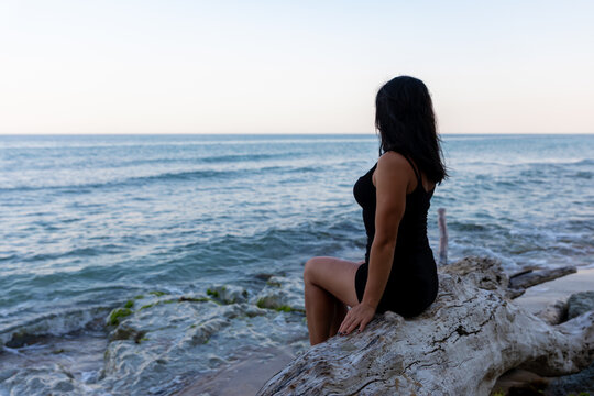 Woman in black dress sits on driftwood, gazing at the tranquil ocean under a clear sky, evoking calm and contemplation.