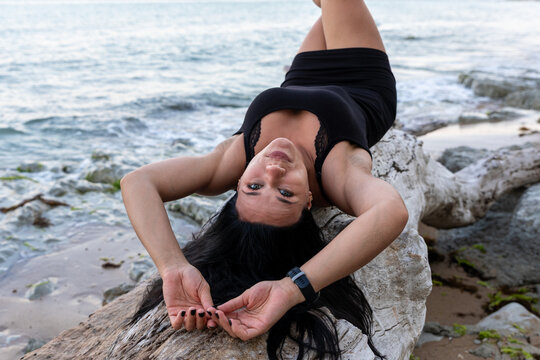 Confident woman in black dress reclining on driftwood by the ocean, enjoying the seaside with waves and natural scenery in the background.