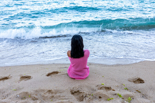 Rear view of woman in pink dress sitting on sandy beach facing ocean waves, evoking solitude and relaxation.