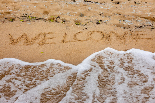 The word 'Welcome' is written in smooth sand as gentle ocean waves approach, ideal for hospitality, travel, and vacation concepts.