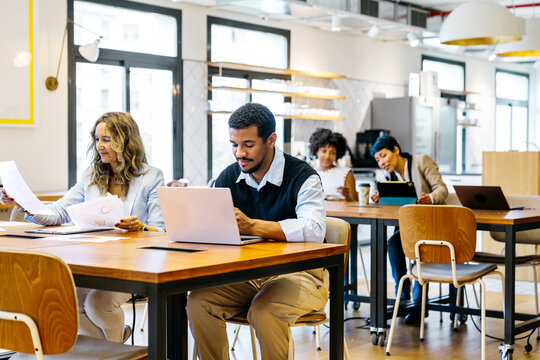 Diverse business workers collaborating and working on laptops, tablets, and documents in a modern, open plan coworking office environment