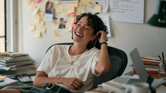 Software developer leans back at a cluttered home office desk while a spontaneous candid moment captures her laughing mid call during a virtual project site meeting, sticky notes