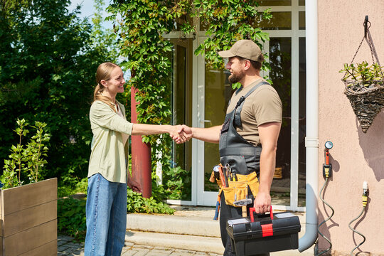 Woman shaking hands with male technician holding toolbox outside house. Image represents customer satisfaction and home repair services, suitable for local business marketing