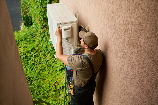 Caucasian young adult male technician repairing outdoor air conditioning unit on building wall. Image represents home maintenance services and professional HVAC repair, high angle view