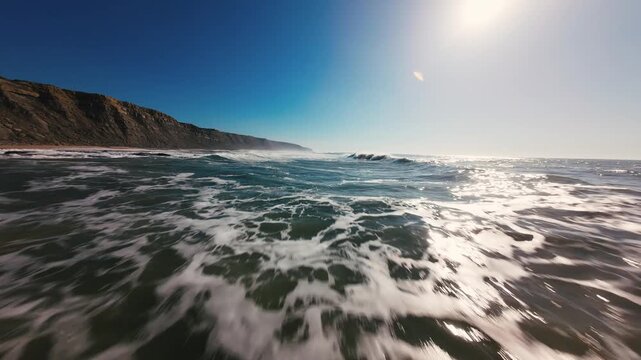 Sun glints on ocean waves near rocky cliffs. Water crashes with white foam under bright sky. View from water level captures dynamic motion. Ideal for travel, adventure, or nature-themed projects