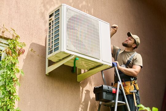 Caucasian young adult male technician standing on ladder repairing outdoor air conditioning unit. Image suitable for home maintenance services and HVAC repair advertising