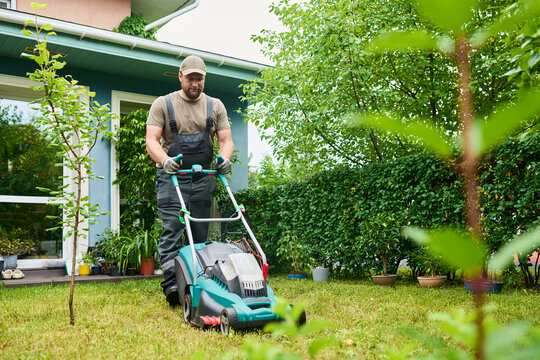 Caucasian man pushing lawn mower in backyard, maintaining green grass. Professional landscaping service concept, ideal for home improvement and gardening promotion