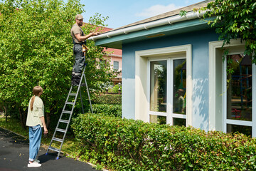 Caucasian male contractor standing on ladder repairing roof gutter while Caucasian female homeowner watches. Image represents home maintenance services and residential property care © pressmaster