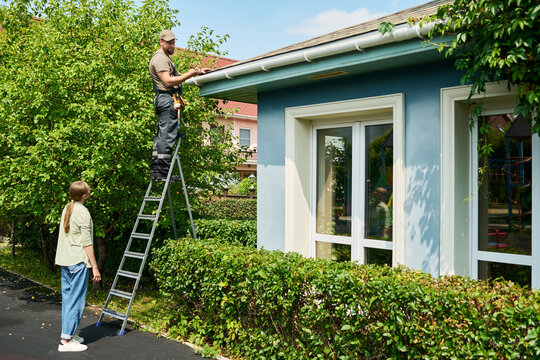 Caucasian male contractor standing on ladder repairing roof gutter while Caucasian female homeowner watches. Image represents home maintenance services and residential property care