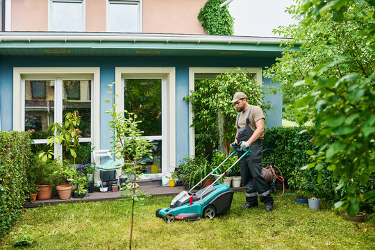 Caucasian young adult male gardener pushing lawn mower in backyard of residential house. Image represents professional landscaping services and home maintenance marketing, wide shot
