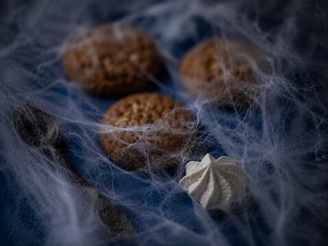 Three oatmeal cookies and a meringue cookie placed on a blue fabric surface covered in artificial spider webs with a vintage metal spoon