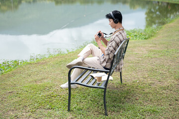 Fototapeta premium Asian man with headphones sitting alone on bench by lake and enjoy photographing the natural scenery