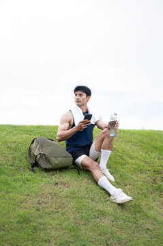 Athletic Asian man resting with phone and water bottle on grass after exercising in the green park.