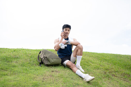 Athletic Asian man talking on phone while sitting on grass hill after a fitness session in the park