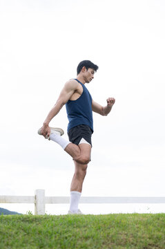 Full body of athletic asian man stretching legs over a grassy field in green park under a bright sky