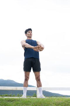 Athletic Asian man standing and stretching arms while enjoying a green landscape before exercise.