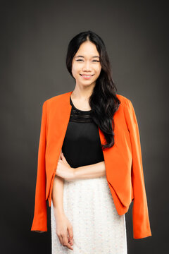 Portrait of a smiling young Southeast Asian Malaysian woman wearing professional corporate attire, featuring a bright orange blazer and black top with a confident pose