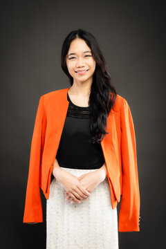 Portrait of a smiling young Southeast Asian Malaysian woman wearing professional corporate attire, featuring a bright orange blazer and black top with a confident pose
