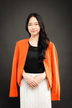Portrait of a smiling young Southeast Asian Malaysian woman wearing professional corporate attire, featuring a bright orange blazer and black top with a confident pose