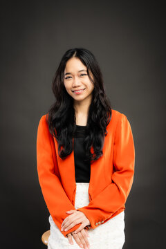 Portrait of a smiling young Southeast Asian Malaysian woman wearing professional corporate attire, featuring a bright orange blazer and black top with a confident pose