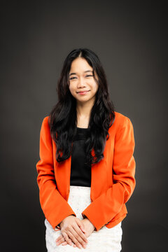 Portrait of a smiling young Southeast Asian Malaysian woman wearing professional corporate attire, featuring a bright orange blazer and black top with a confident pose