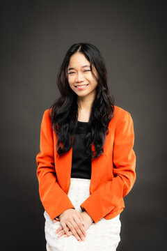 Portrait of a smiling young Southeast Asian Malaysian woman wearing professional corporate attire, featuring a bright orange blazer and black top with a confident pose