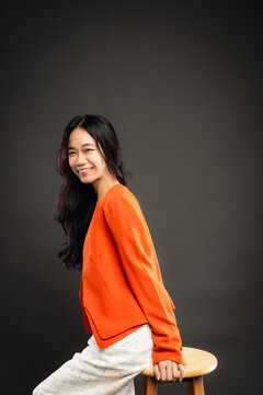Portrait of a smiling young Southeast Asian Malaysian woman wearing professional corporate attire, featuring a bright orange blazer and black top with a confident pose