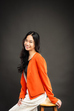 Portrait of a smiling young Southeast Asian Malaysian woman wearing professional corporate attire, featuring a bright orange blazer and black top with a confident pose