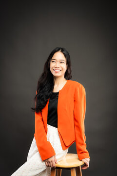 Portrait of a smiling young Southeast Asian Malaysian woman wearing professional corporate attire, featuring a bright orange blazer and black top with a confident pose